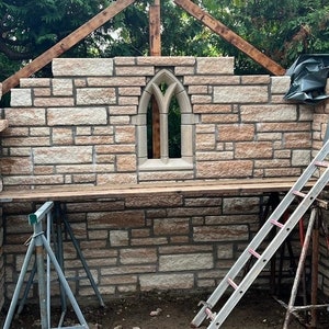 May include: A stone wall with a pointed arch window, a wooden frame above, and two ladders on the ground. The wall is made of light brown and beige stones.