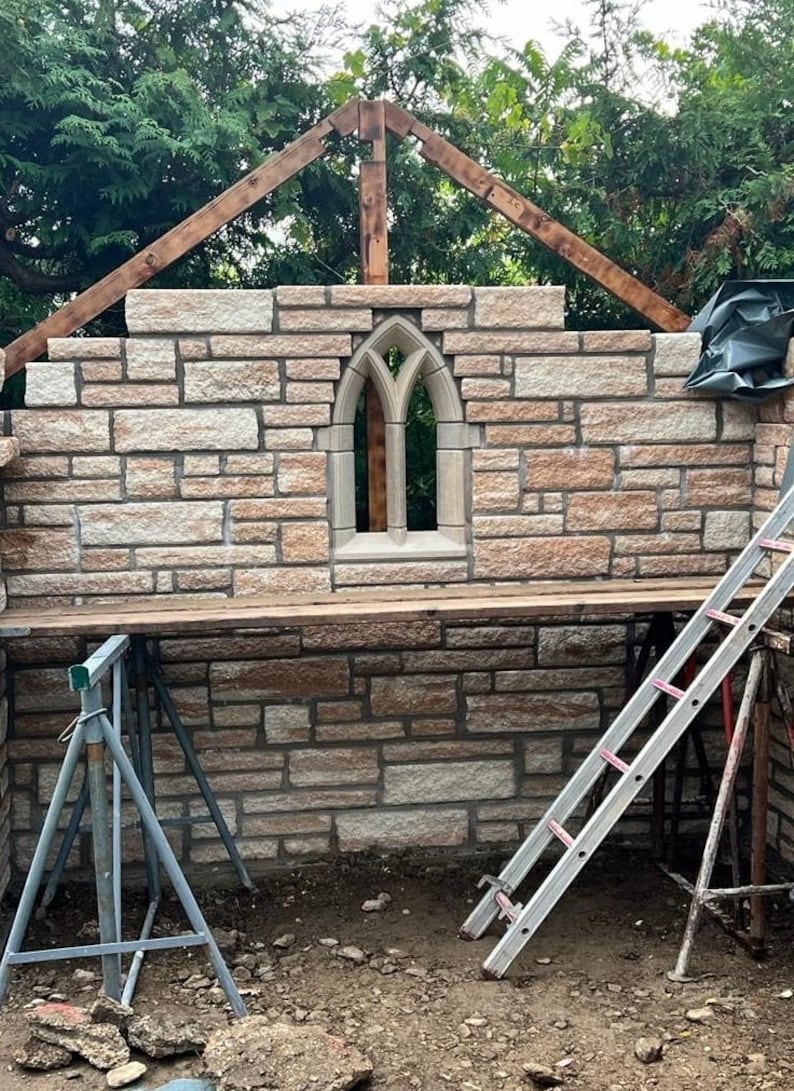 May include: A stone wall with a pointed arch window, a wooden frame above, and two ladders on the ground. The wall is made of light brown and beige stones.