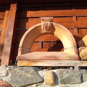 May include: A terracotta-colored, arched architectural element with a decorative top. The arch is supported by a rectangular base and features carved leaf details. The structure is set against a wooden fence and stone wall.