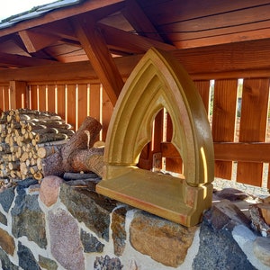 May include: A tan-colored stone window frame featuring a pointed gothic arch design with a flat base, positioned upright on a stone wall surface. The architectural element has a smooth, matte finish and a symmetrical, arched opening.