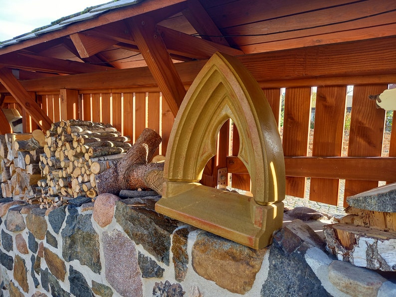 May include: A tan-colored stone window frame featuring a pointed gothic arch design with a flat base, positioned upright on a stone wall surface. The architectural element has a smooth, matte finish and a symmetrical, arched opening.