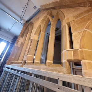 May include: A close-up of a stone archway with multiple arched openings. The archway is made of light brown stone and has a rough texture. The archway is illuminated by natural light, casting shadows on the stone.