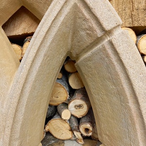 May include: A close-up of a stone archway with stacked logs of firewood inside. The archway is made of rough-textured concrete and has a light brown color.