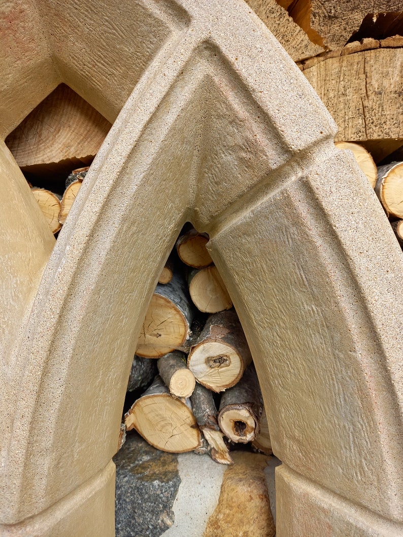 May include: A close-up of a stone archway with stacked logs of firewood inside. The archway is made of rough-textured concrete and has a light brown color.