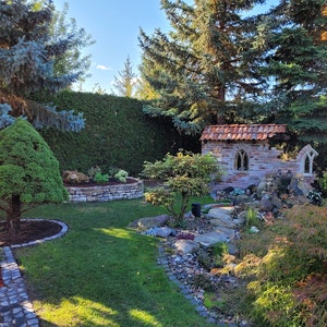 May include: A small brick building with a red tile roof and arched windows sits in a garden setting. A stone path leads to the building, and there are trees and shrubs surrounding it.