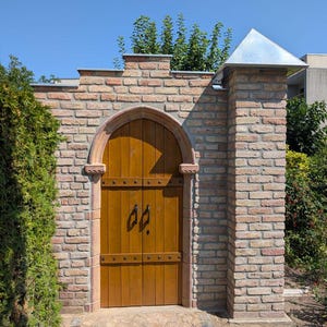 May include: A double wooden door with a pointed arch top set into a light brown brick wall. The door features black metal handles and horizontal metal bands. A brick pillar with a silver metal pyramid-shaped cap stands to the right side.