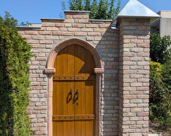 May include: A double wooden door with a pointed arch top set into a light brown brick wall. The door features black metal handles and horizontal metal bands. A brick pillar with a silver metal pyramid-shaped cap stands to the right side.