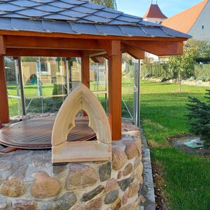 May include: A light-colored stone window frame with a pointed gothic arch sits on a circular stone well structure. The well features a wooden roof supported by brown timber posts and a circular wooden platform inside the stone base.