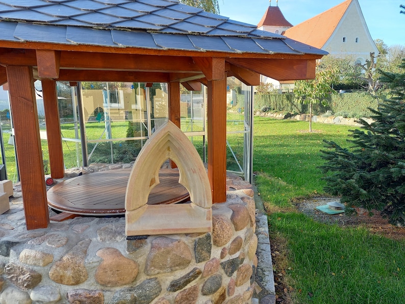 May include: A light-colored stone window frame with a pointed gothic arch sits on a circular stone well structure. The well features a wooden roof supported by brown timber posts and a circular wooden platform inside the stone base.