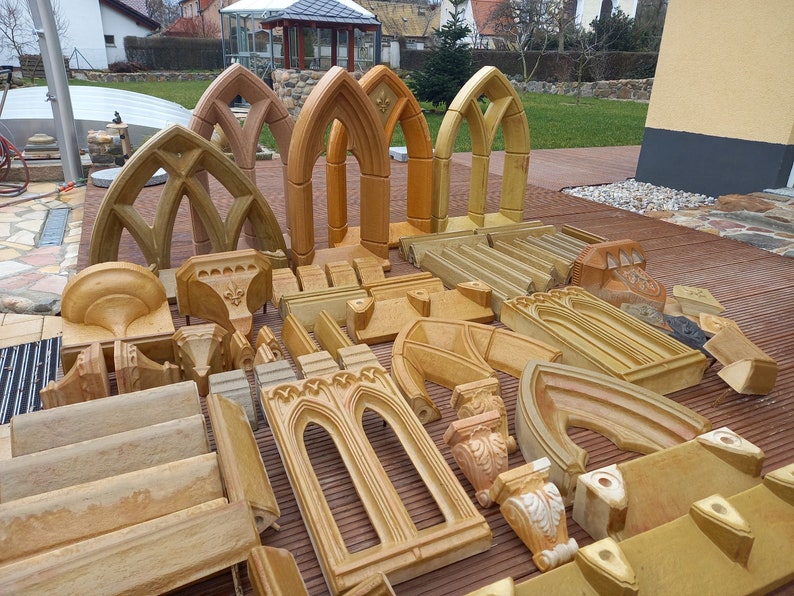 May include: A collection of decorative architectural elements made of light brown stone. The pieces include arches, moldings, and corbels. The pieces are arranged on a wooden deck.