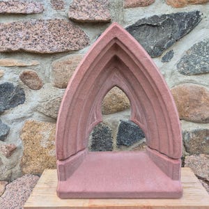 May include: A reddish-brown stone sculpture featuring a pointed gothic archway design. The structure consists of a flat rectangular base supporting a tall, arched frame with recessed molding details, creating an open, hollow center.