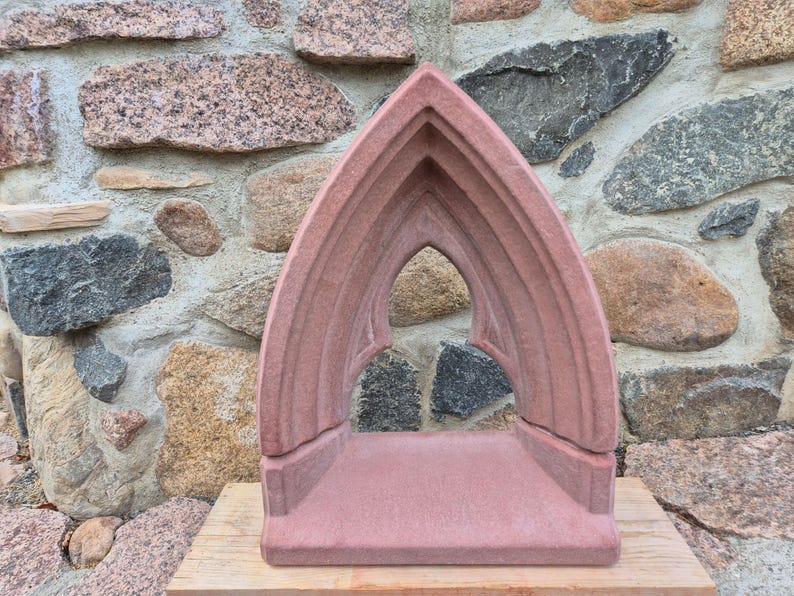 May include: A reddish-brown stone sculpture featuring a pointed gothic archway design. The structure consists of a flat rectangular base supporting a tall, arched frame with recessed molding details, creating an open, hollow center.