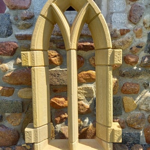 Könnte beinhalten: Ein dekorativer Stein-Fensterrahmen mit einem Spitzbogen-Design. Der Rahmen ist aus hellem braunem Stein gefertigt und hat eine raue Textur. Er steht auf einer Holzplattform.