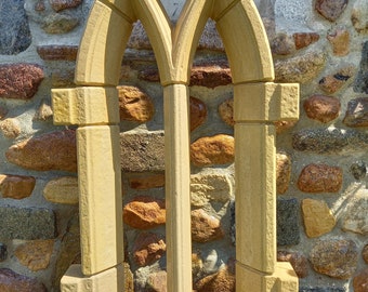 May include: A decorative stone window frame with a pointed arch design. The frame is made of light brown stone and has a rough texture. It is sitting on a wooden platform.
