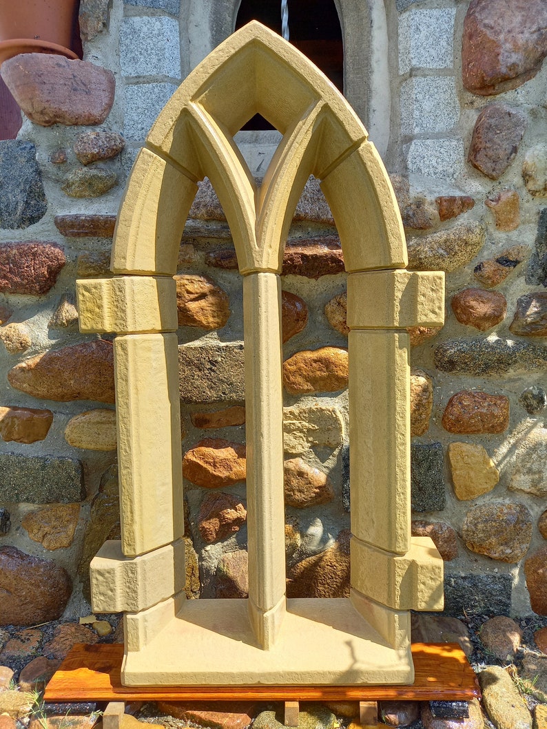 May include: A decorative stone window frame with a pointed arch design. The frame is made of light brown stone and has a rough texture. It is sitting on a wooden platform.