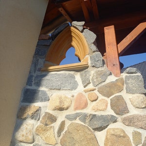 May include: A stone wall featuring a pointed arch window frame made of light-colored wood. The wall is constructed from irregularly shaped gray, tan, and reddish stones set in mortar, positioned beneath a wooden roof structure against a blue sky.