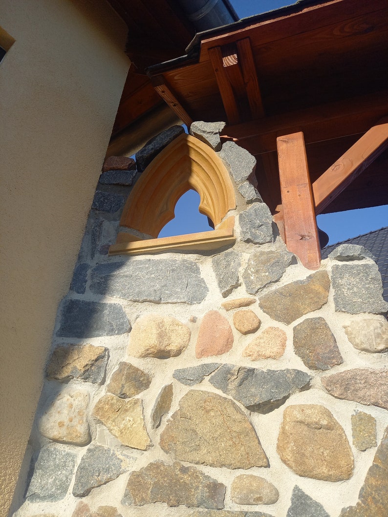May include: A stone wall featuring a pointed arch window frame made of light-colored wood. The wall is constructed from irregularly shaped gray, tan, and reddish stones set in mortar, positioned beneath a wooden roof structure against a blue sky.