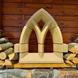 Könnte beinhalten: Ein dekorativer Steinbogen mit einer spitzen Oberseite und zwei gewölbten Öffnungen. Der Bogen ist aus hellem beigefarbenem Stein gefertigt und hat eine raue Textur. Er steht auf einer Holzoberfläche.