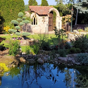 May include: A brick archway with a wooden door leads to a small brick building with a tiled roof. A pond with clear blue water is in the foreground, surrounded by lush green plants and rocks.
