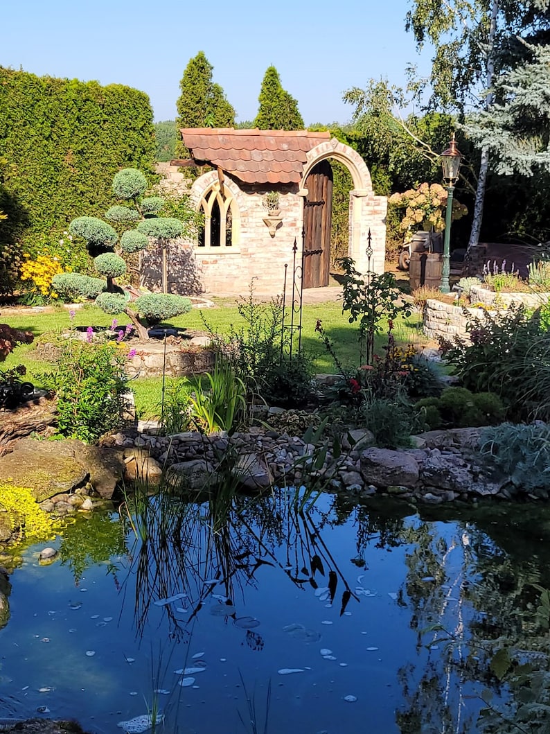 May include: A brick archway with a wooden door leads to a small brick building with a tiled roof. A pond with clear blue water is in the foreground, surrounded by lush green plants and rocks.
