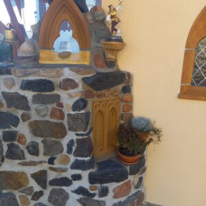 May include: A stone masonry structure featuring a pointed arch window frame, a small figurine of a person in gold and brown clothing, a recessed arched niche, and two potted plants with green and reddish foliage set against a yellow wall.