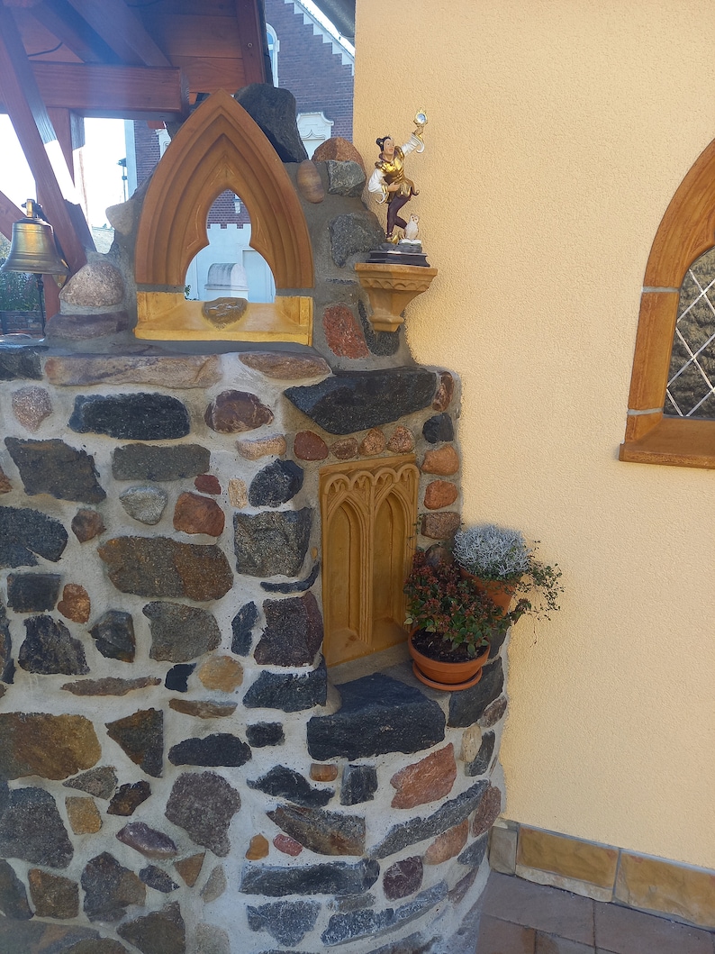 May include: A stone masonry structure featuring a pointed arch window frame, a small figurine of a person in gold and brown clothing, a recessed arched niche, and two potted plants with green and reddish foliage set against a yellow wall.