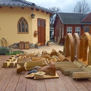 May include: A collection of unfinished, light brown, wooden archway mouldings. The mouldings are arranged on a wooden deck in front of a yellow building with a brown door.