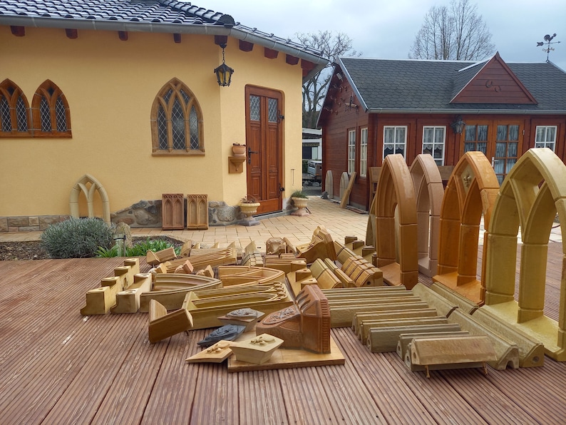 May include: A collection of unfinished, light brown, wooden archway mouldings. The mouldings are arranged on a wooden deck in front of a yellow building with a brown door.