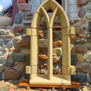 May include: A decorative stone window frame with a pointed arch design. The frame is made of a light brown stone and has a rustic look. It is sitting on a wooden base.