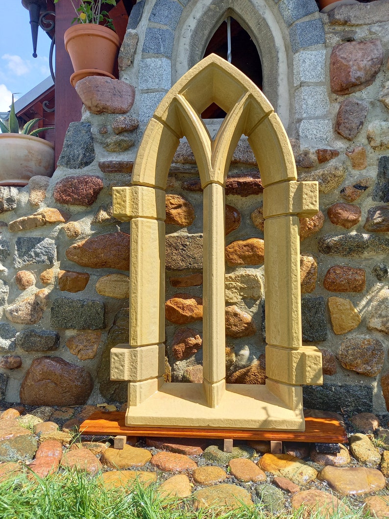 May include: A decorative stone window frame with a pointed arch design. The frame is made of a light brown stone and has a rustic look. It is sitting on a wooden base.