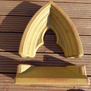 May include: A two-piece mustard yellow stone-like object consisting of a pointed arch frame and a rectangular base. The arch features tiered molding and a sharp apex, while the base has a flat top and angled sides with a textured finish.
