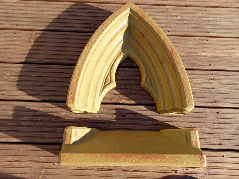 May include: A two-piece mustard yellow stone-like object consisting of a pointed arch frame and a rectangular base. The arch features tiered molding and a sharp apex, while the base has a flat top and angled sides with a textured finish.