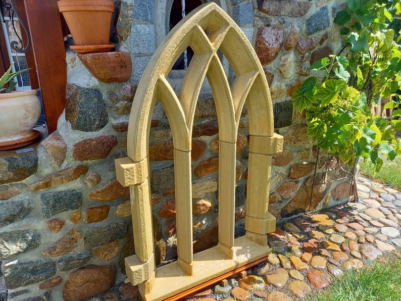 May include: A decorative stone archway with a gothic design. The archway is made of light brown stone and has three arched openings. The archway is standing in front of a stone wall.