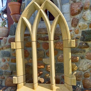May include: A decorative stone window frame with a pointed arch design. The frame is made of light beige stone and has three arched openings. The frame is sitting on a wooden base.
