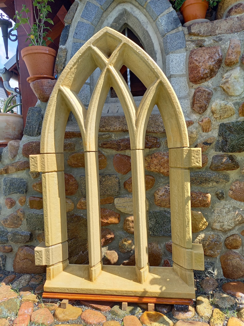 May include: A decorative stone window frame with a pointed arch design. The frame is made of light beige stone and has three arched openings. The frame is sitting on a wooden base.