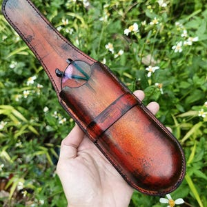 May include: A handcrafted, brown leather glasses case with a rich, distressed finish. The case is designed to hold a pair of glasses, with a secure flap closure. The case is held in a hand, showcasing the craftsmanship and the glasses inside.