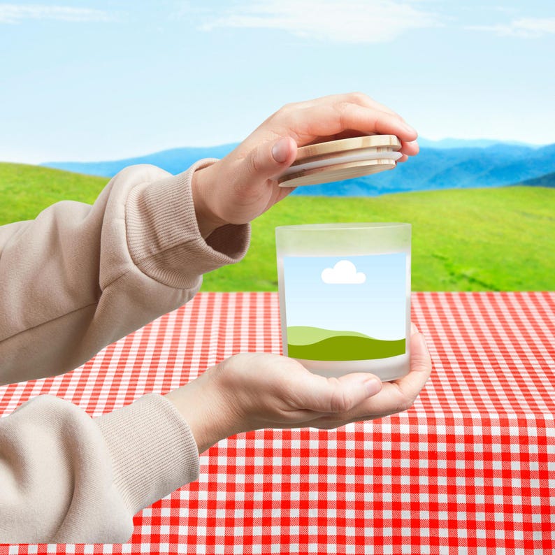 Peut inclure: Un pot &agrave; bougie en verre d&eacute;poli avec un couvercle en bois. Le pot pr&eacute;sente un paysage avec un ciel bleu, un nuage blanc et des collines vertes. Le pot est tenu au-dessus d'une nappe &agrave; carreaux rouges et blancs.