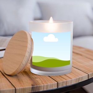 Puede incluir: Una vela de vidrio esmerilado encendida con una tapa de madera sobre una mesa de madera. La vela tiene una etiqueta con un diseño de cielo azul, nube blanca y colinas verdes. La tapa está al lado de la vela.