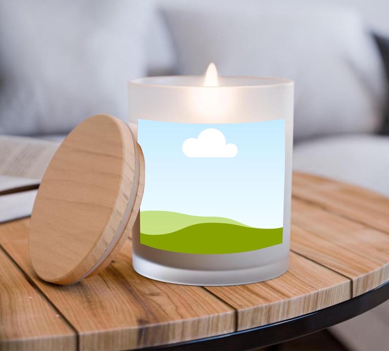 Peut inclure: Une bougie en verre d&eacute;poli allum&eacute;e avec un couvercle en bois sur une table en bois. La bougie a une &eacute;tiquette avec un ciel bleu, un nuage blanc et un motif de collines vertes. Le couvercle est &agrave; c&ocirc;t&eacute; de la bougie.