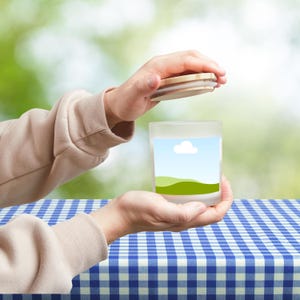 Peut inclure: Un pot en verre d&eacute;poli avec un couvercle en bois, ouvert. Le pot pr&eacute;sente un paysage avec un ciel bleu, un nuage blanc et des collines vertes. Le pot est tenu au-dessus d'une nappe &agrave; carreaux bleue et blanche.