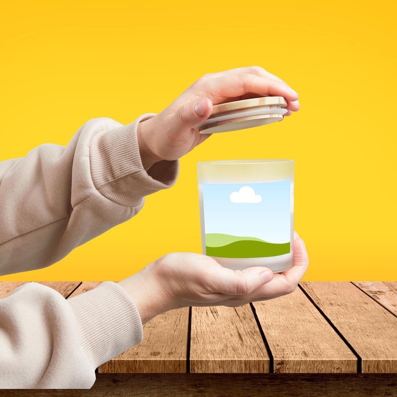 Peut inclure: Un pot &agrave; bougie en verre d&eacute;poli avec un couvercle en bois. Le pot pr&eacute;sente un paysage avec un ciel bleu, un nuage blanc et des collines vertes. Le pot est tenu dans les mains d'une personne sur un fond jaune.