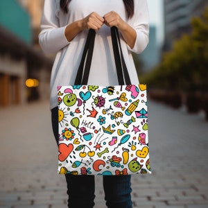 May include: A white tote bag with black handles features a colorful doodle pattern. The pattern includes a sun, a heart, a crown, a ladybug, a fish, a star, a bird, a bone, a butterfly, a sock, a cup, a balloon, a cloud, a cherry, a rainbow, and the word "Hello".
