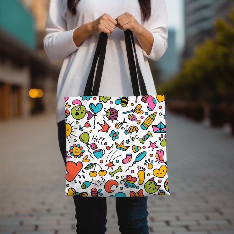 May include: A white tote bag with black handles features a colorful doodle pattern. The pattern includes a sun, a heart, a crown, a ladybug, a fish, a star, a bird, a bone, a butterfly, a sock, a cup, a balloon, a cloud, a cherry, a rainbow, and the word "Hello".