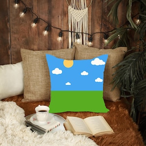 Puede incluir: Cojín cuadrado con un diseño de cielo azul y campo verde. El cojín presenta un sol amarillo y nubes blancas. El cojín está colocado sobre una superficie marrón con otros cojines y objetos decorativos.