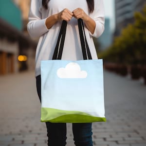 May include: A tote bag with black straps and a sky blue background. The bag has a white cloud and green hills printed on it.