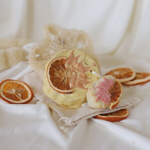 May include: A white and pink soap bar with a dried orange slice on top. The soap bar is sitting on a white cloth with a natural sponge and other dried orange slices.