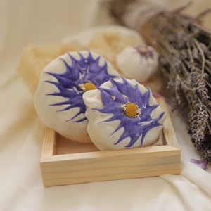 Pode incluir: Duas bombas de banho brancas com desenhos de flores roxas e amarelas em uma bandeja de madeira. As bombas de banho são cercadas por lavanda seca.