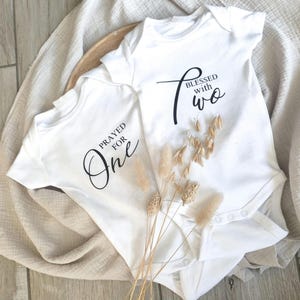 Könnte beinhalten: Zwei weiße Baby-Bodys mit schwarzer Schrift. Einer trägt die Aufschrift "Prayed for One", der andere "Blessed with Two". Die Bodys liegen auf einer neutralfarbenen Oberfläche mit getrockneten Blumen.