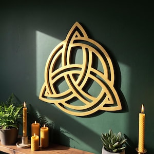 May include: A gold-coloured triquetra wall decoration featuring an intricate, interlaced knot design. The piece is constructed with smooth, curved lines that form a continuous triangular shape, displaying a metallic finish that reflects light.
