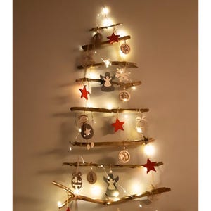 Può includere: Una struttura per albero di Natale da parete composta da rami di legno galleggiante orizzontali disposti a forma di triangolo. I rami sono decorati con ornamenti in legno appesi, tra cui stelle rosse, angeli e motivi circolari, illuminati da luci a corda bianco caldo.